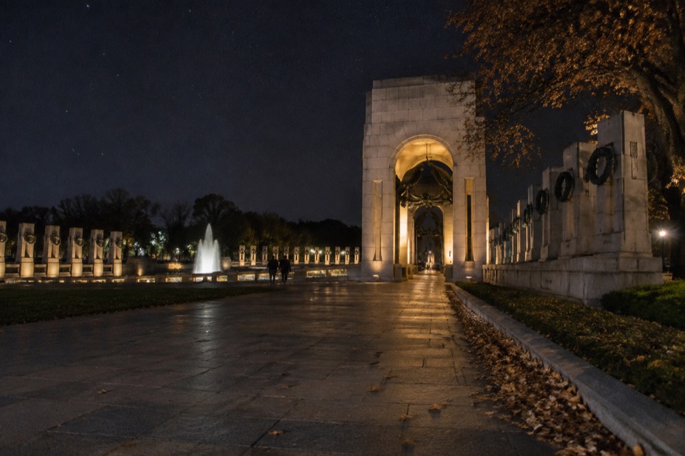 A stone memorial arch lit by warm floodlights beside an empty paved path at night