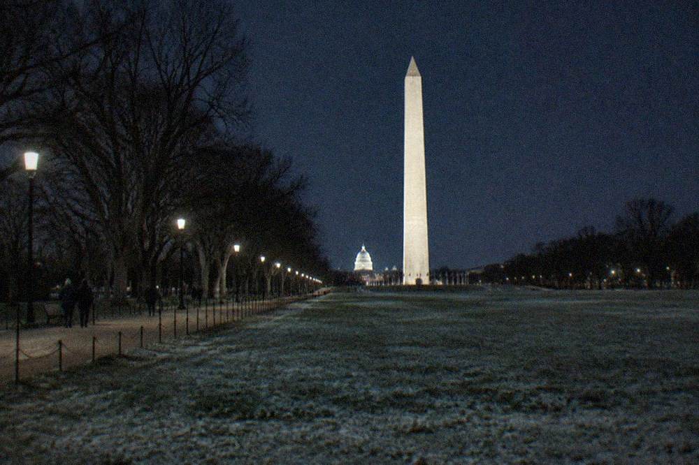 The Washington Monument illuminated at night, a tall obelisk glowing against a deep blue sky with the Capitol dome visible in the distance