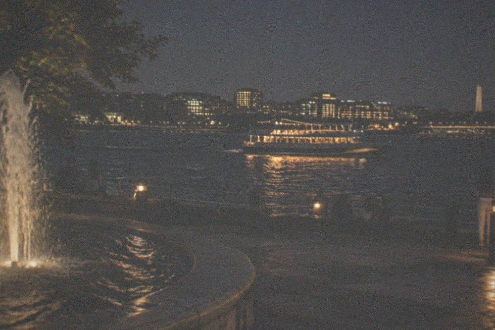 A dinner boat lit up and underway on the Potomac River with the city skyline behind it