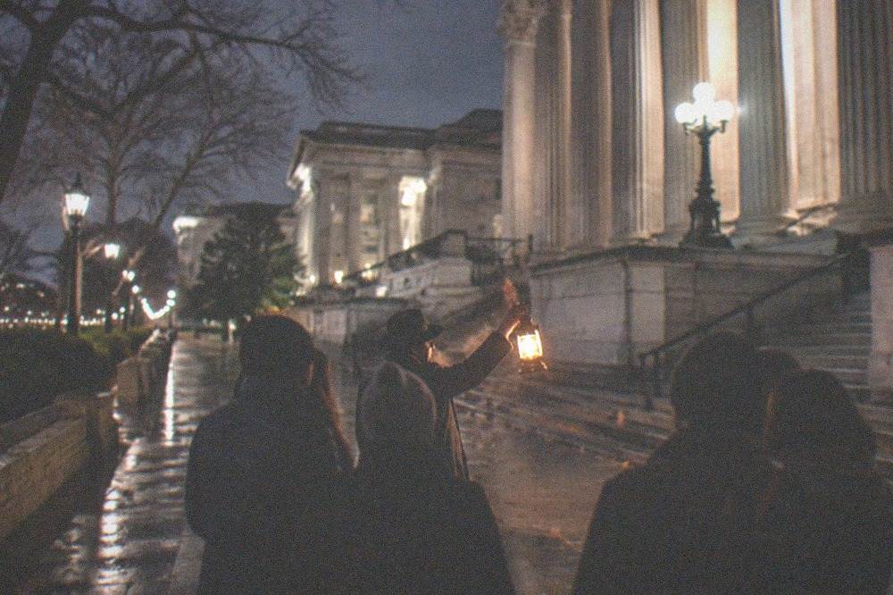 A guide holding a lantern addressing a small group on a dark city sidewalk