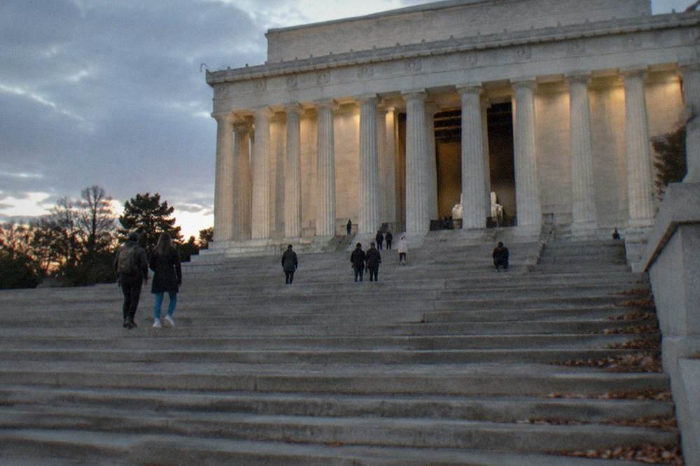 The illuminated Lincoln Memorial steps and columns under a dark evening sky with a few visitors on the stairs