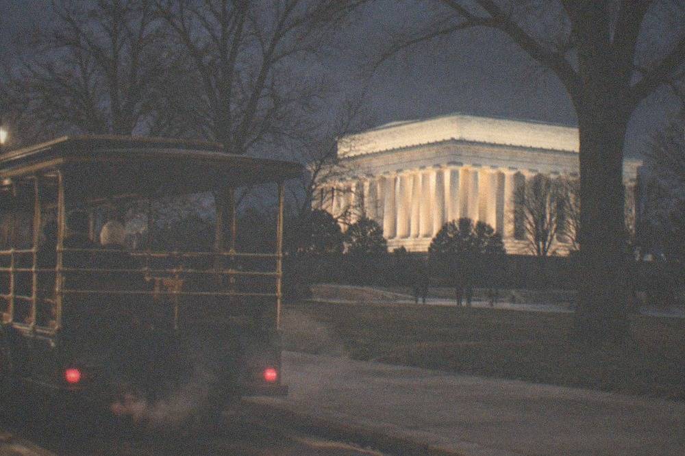 An illuminated open tour trolley parked near the National Mall with lit memorials in the background at night