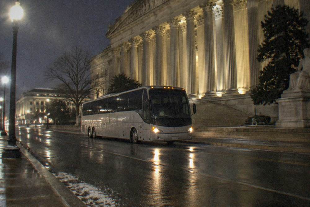 A lit tour bus parked near the glowing white columns of a memorial entrance at night