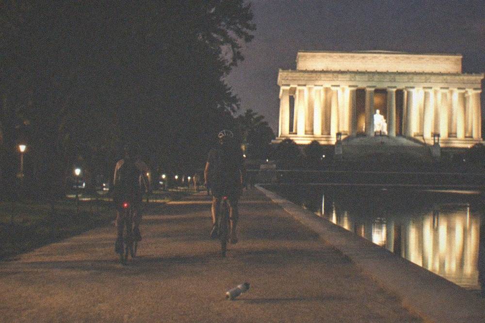 Cyclists riding past floodlit memorial columns on an empty Mall pathway at night