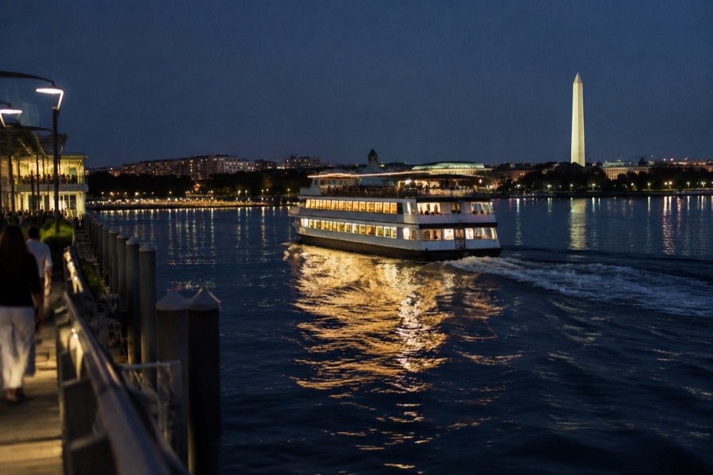 A buffet dinner cruise boat on the Potomac River at night with the illuminated city skyline behind it