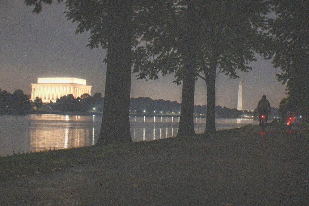 Cyclists with lights riding along a tree-lined path near lit memorials at night
