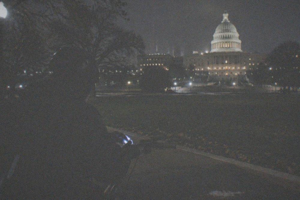 A cyclist in a light jacket and helmet, adjusting a bike light before a night ride with the Capitol dome visible in the distance.