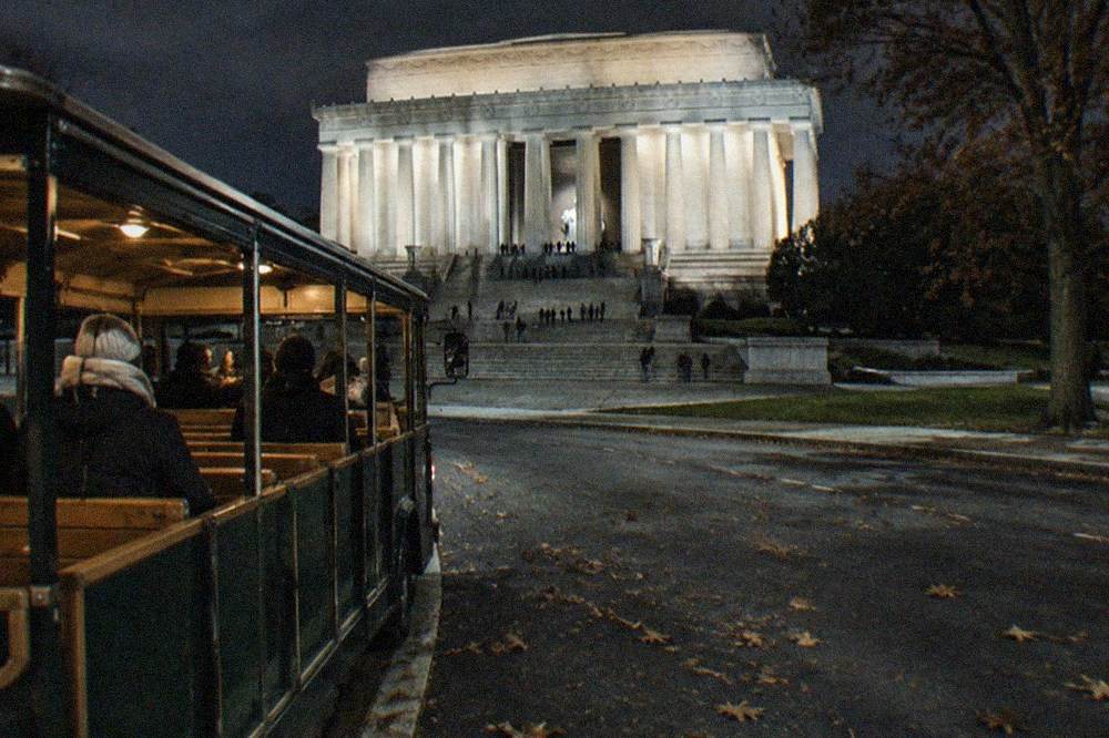 An open tour trolley parked beside illuminated memorial columns at night